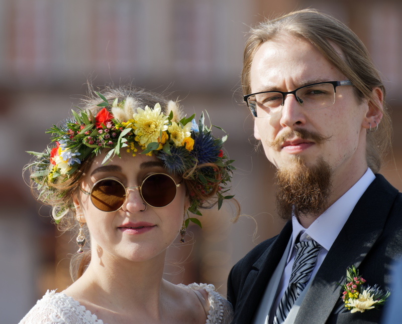 3 - A couple in wedding attire, featuring a person wearing a white lace top with a vibrant floral crown and round sunglasses alongside a partner in a classic black suit and patterned tie
